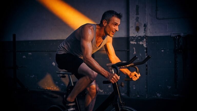 Adulto de unos 45 años pedaleando en bicicleta estática durante un intervalo de alta intensidad en gimnasio industrial minimalista, iluminación amber cenital con sombra dramática sobre fondo navy-black vignette.