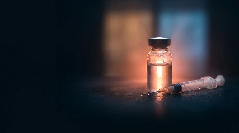 Un vial de solución peptídica con brillo coral y una jeringa fina sobre fondo oscuro dividido en dos zonas de luz, que representa el doble estatus regulatorio de la tesamorelina.