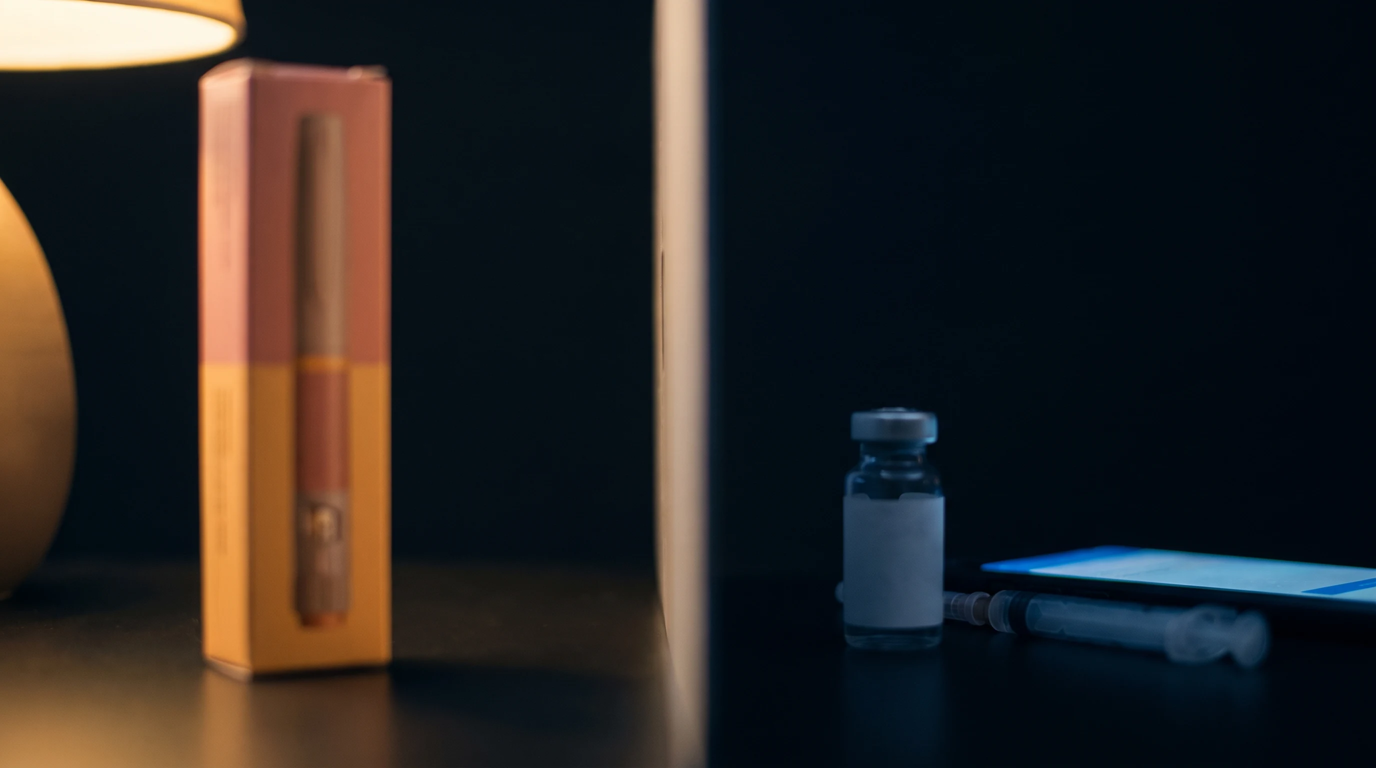 A la izquierda, una caja de inyectable sellada bajo luz c&aacute;lida de farmacia; a la derecha, un vial suelto sin etiqueta y una jeringa junto al brillo de un tel&eacute;fono, representando el mercado gris.